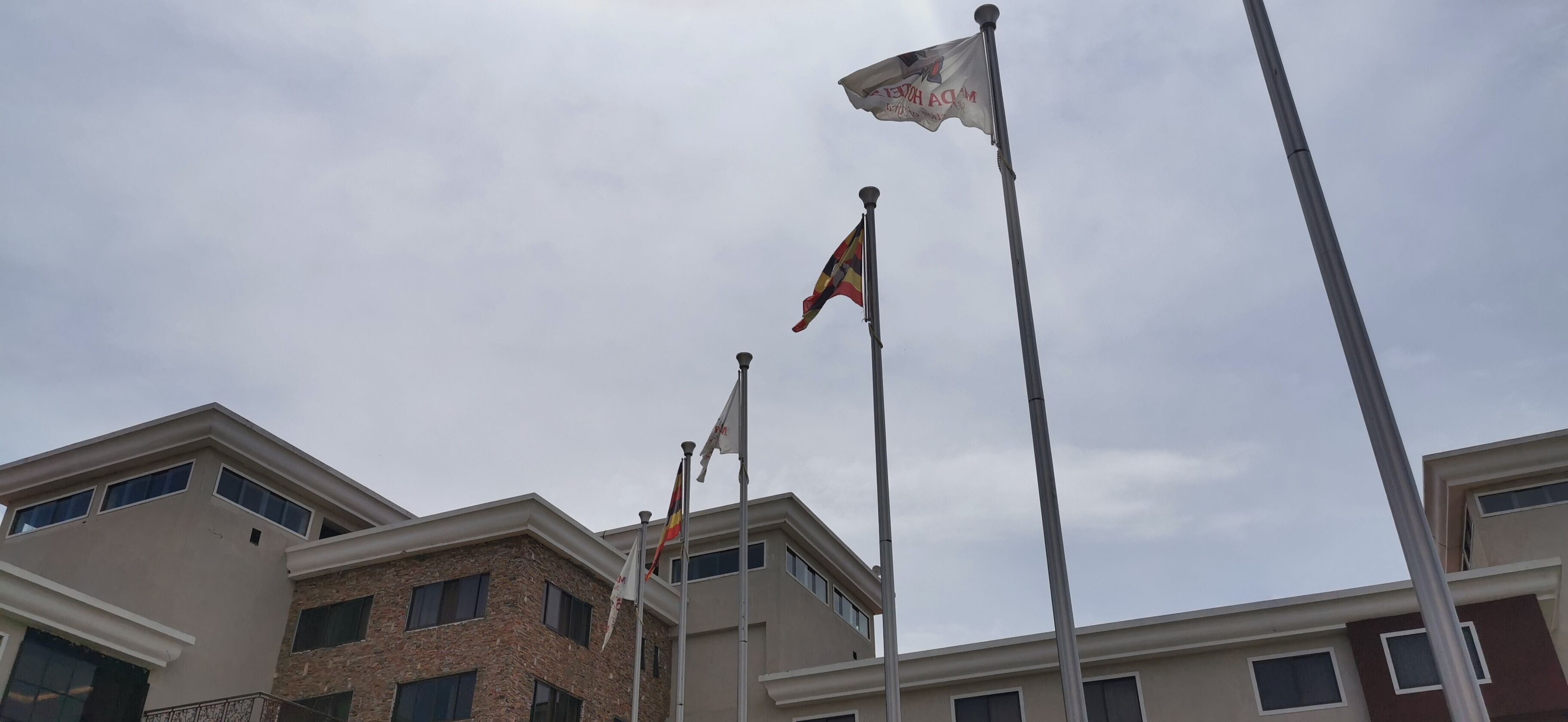 A row of flagpoles with flags, set against a cloudy sky, in front of a building with multiple windows and a flat roof.