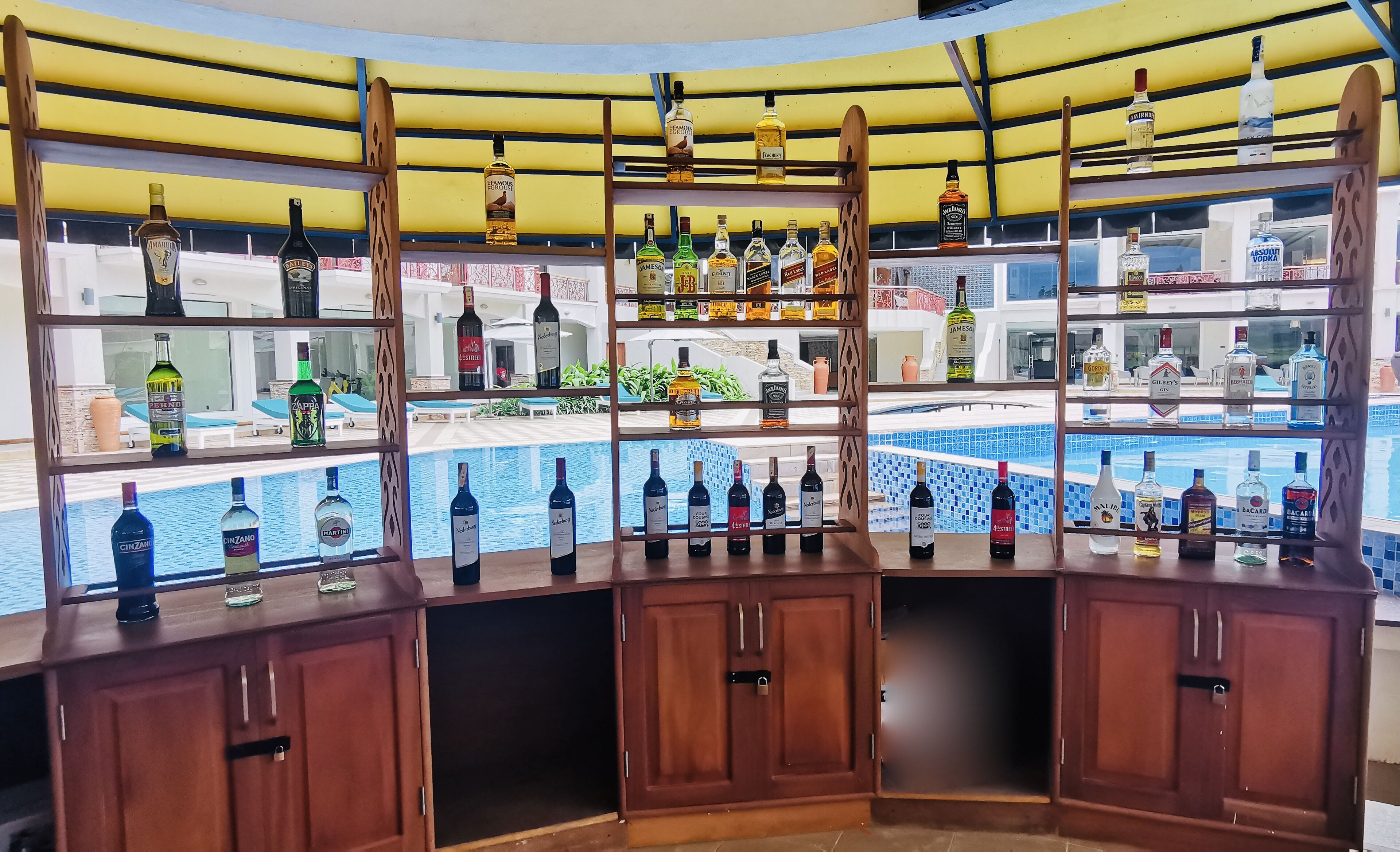 A bar area with shelves of various liquor bottles, a pool view, and wooden cabinets.