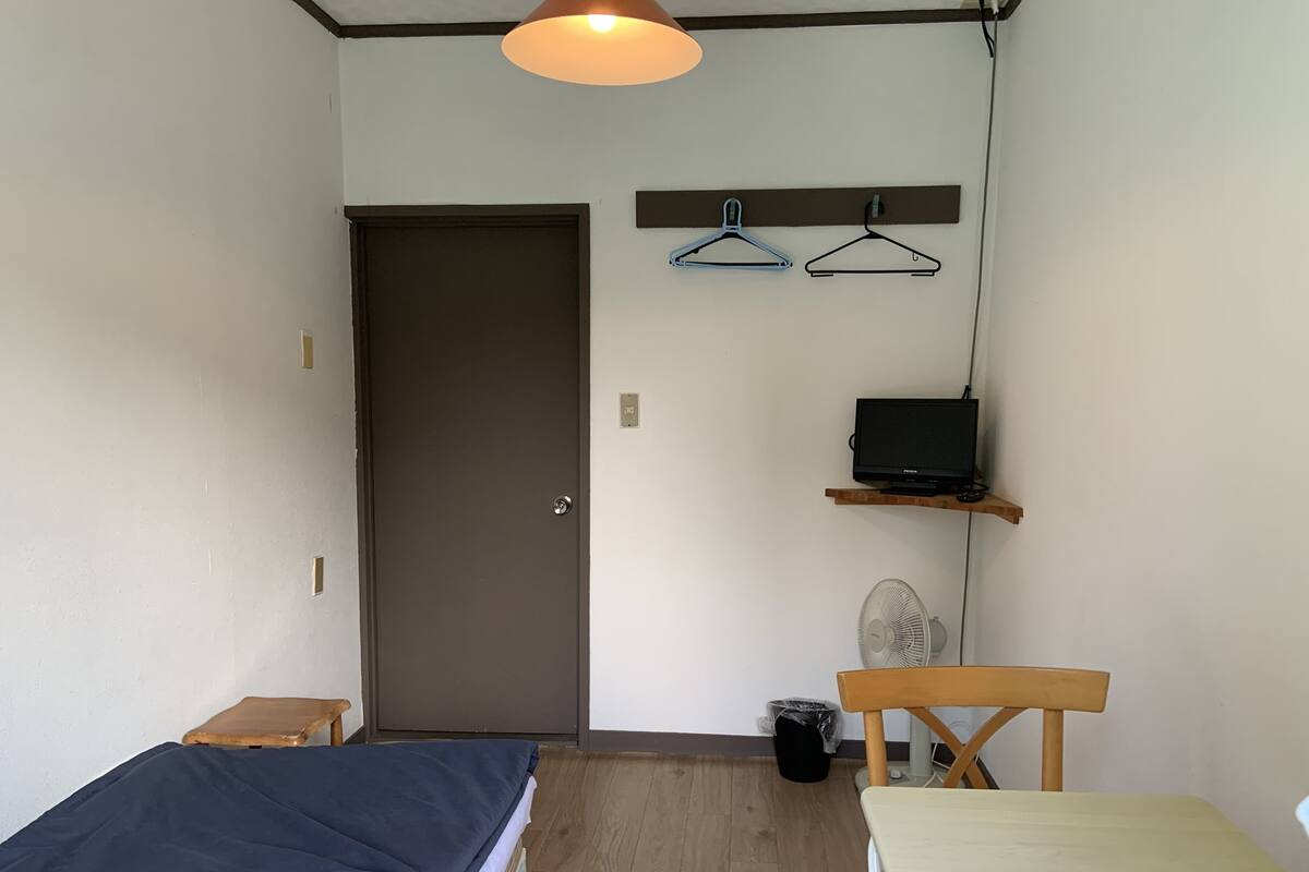 A small, well-lit room with a bed, a table, a chair, a desk with a computer, a fan, and a door.