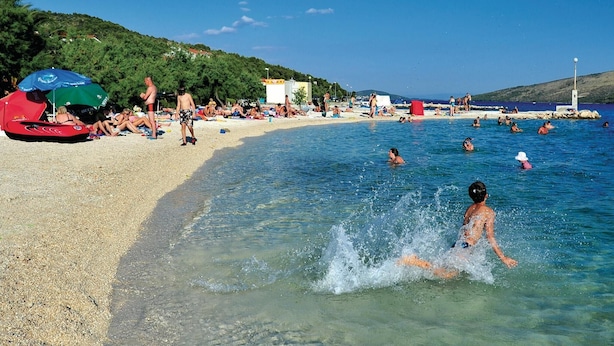 Una spiaggia con acque cristalline, persone che nuotano e si rilassano al sole, e un ombrellone rosso sulla sabbia.