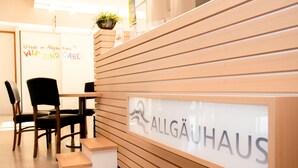 A reception desk with a sign reading "ALLGÄUHAUS" and a plant on top. There are wooden chairs and a small table in front of the desk. - Allgäuhaus Wertach (Wertach)