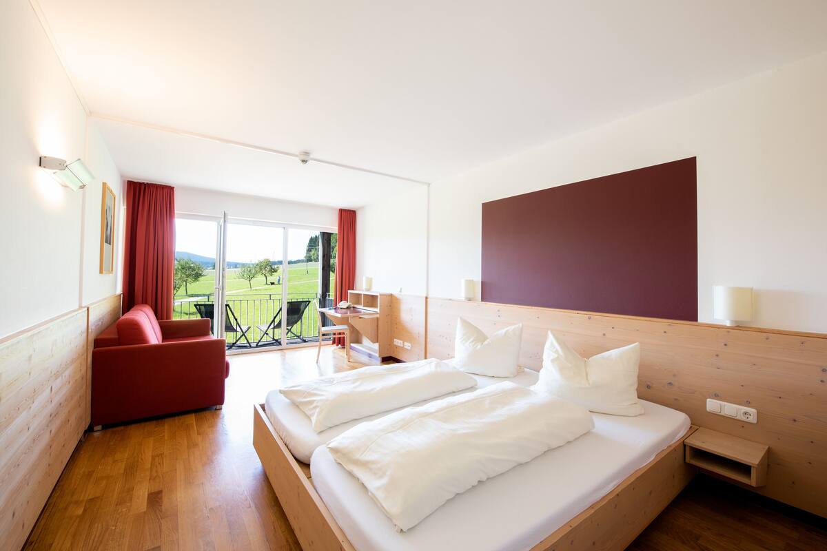 A hotel room with a large bed, a red sofa, a desk, and a view of a green landscape through the window.
