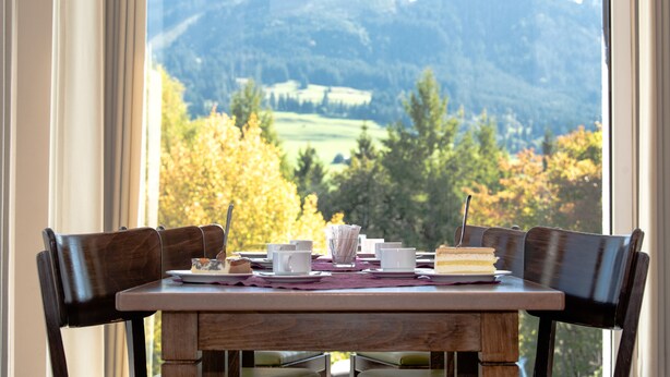 A dining table with chairs, a dessert plate, cups, and a book, set against a window with a view of a mountainous landscape. - Allgäuhaus Wertach (Wertach)