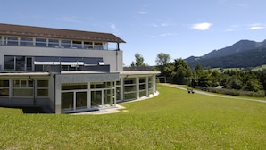 A modern building with large windows and a flat roof, surrounded by a grassy area and mountains in the background. - Allgäuhaus Wertach (Wertach)