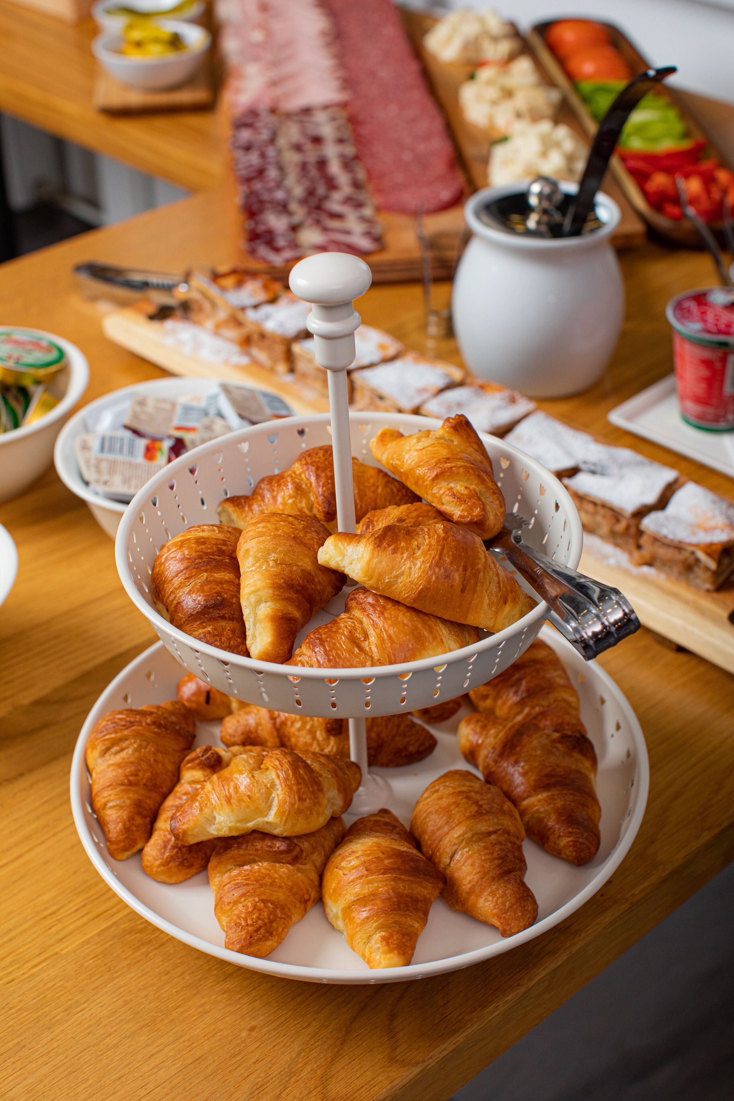 A table with a three-tiered stand holding croissants, a coffee pot, and a variety of other food items.