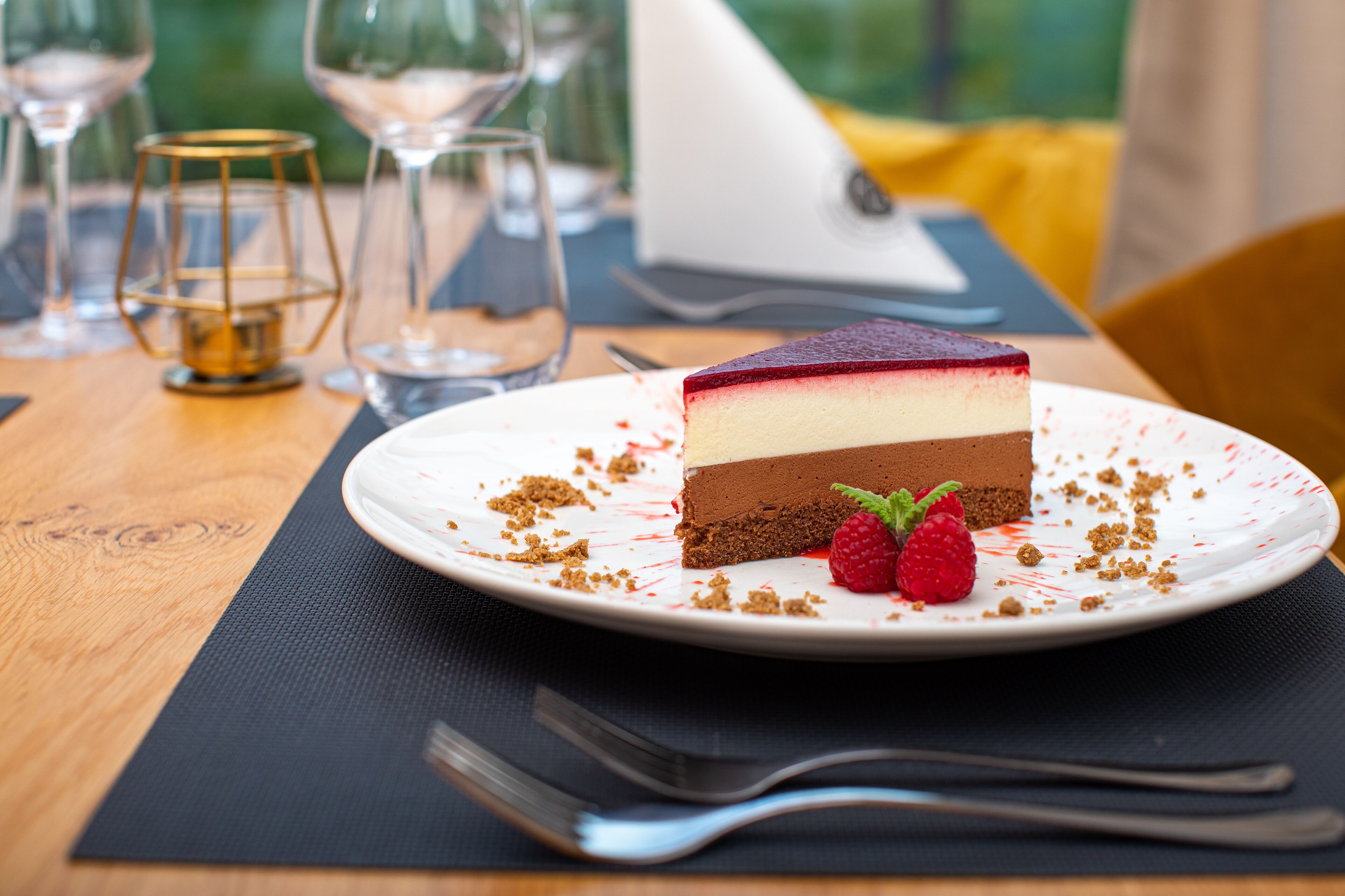 Una rebanada de pastel de queso y chocolate en capas sobre un plato blanco, con frambuesas, acompañada de un tenedor y una copa de vino sobre una mesa de madera.