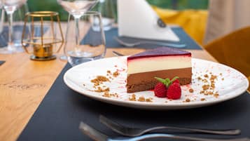 Una rebanada de pastel de queso y chocolate en capas sobre un plato blanco, con frambuesas, acompañada de un tenedor y una copa de vino sobre una mesa de madera.