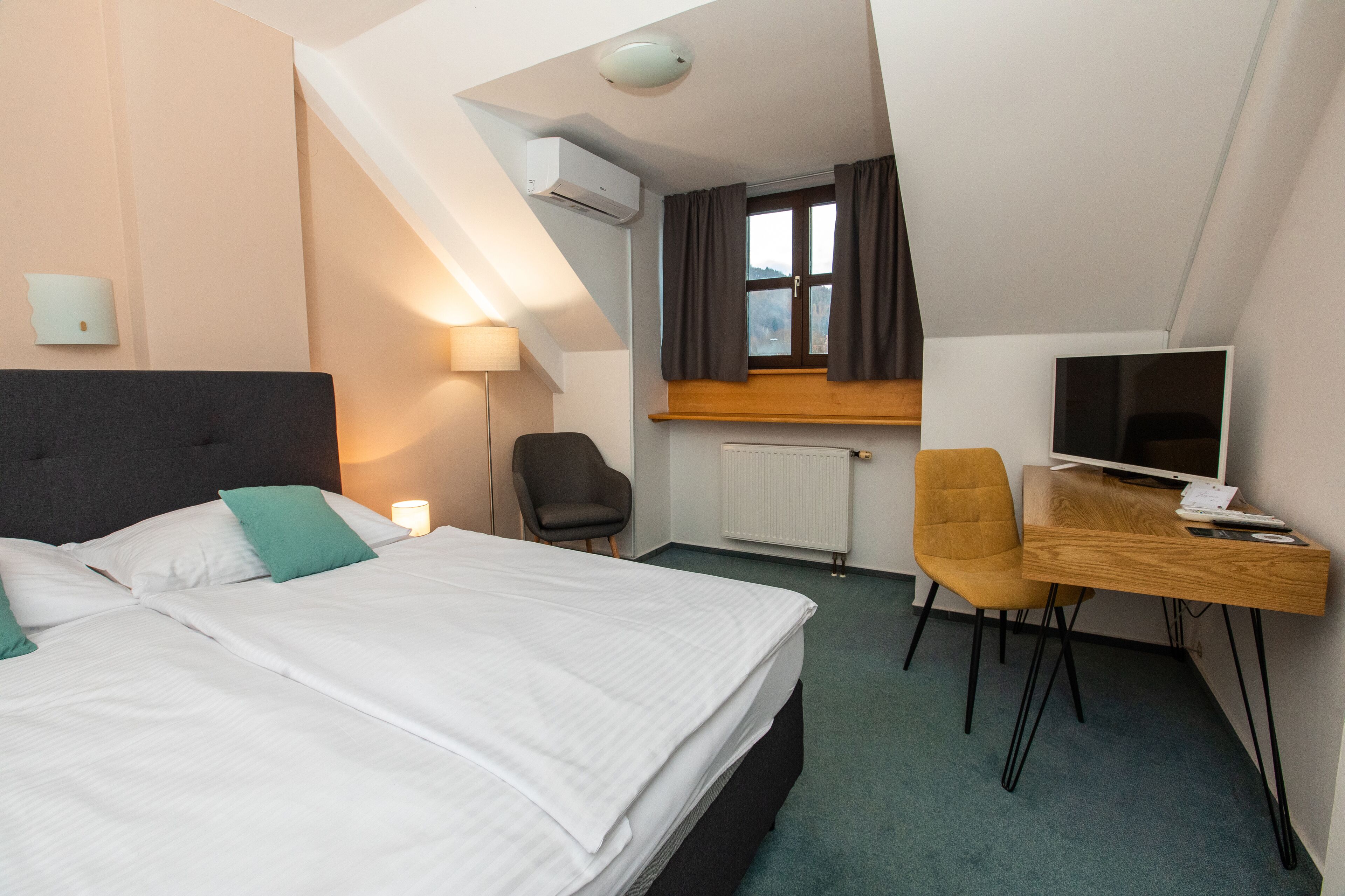 A hotel room with a bed, two chairs, a desk, a TV, and a window with curtains.