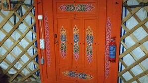 A room with a wooden lattice wall, a red door with decorative patterns, a black mat at the entrance, a fire extinguisher, and a trash bin. - Haller Wooden Houses - Campsite (Budapest)