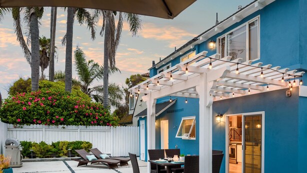 Un patio con mobiliario exterior, una pérgola con luces colgantes y vistas a una casa azul con cerca blanca y plantas florecientes.