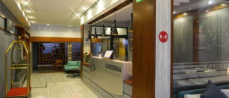 A hotel reception area with a reception desk, a seating area with a sofa and armchairs, a staircase, and a decorative plant.