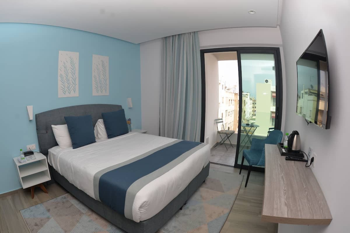 Une chambre d’hôtel moderne dotée d’un grand lit, d’un petit bureau, d’une chaise, d’une télévision et d’un balcon offrant une vue sur la ville.