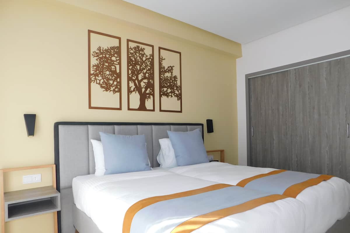 Un lit double avec une tête de lit grise, deux tables de chevet et une petite table de nuit. Trois œuvres d’art sont accrochées au mur et représentent des arbres.
