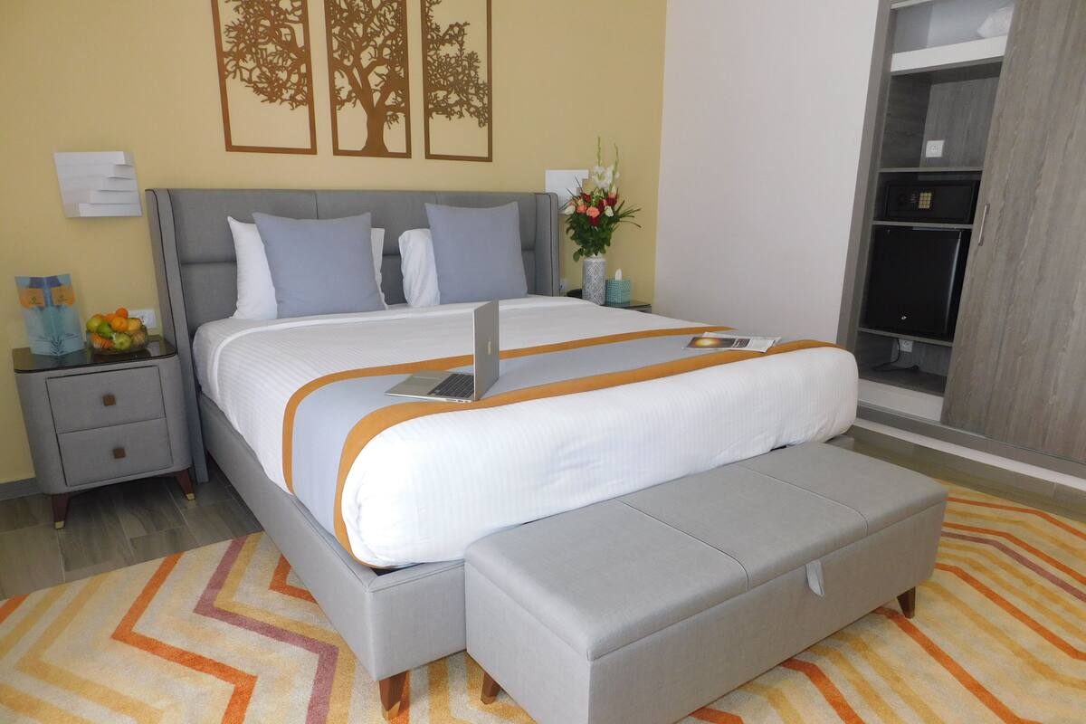 Une chambre moderne avec un grand lit, une tête de lit rembourrée de couleur grise et un pouf assorti. On aperçoit une table de chevet avec un bol de fruits et une lampe. Une commode avec un miroir et un téléviseur se trouve à l’arrière-plan.
