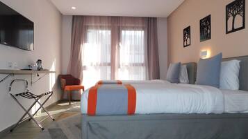 A hotel room with a bed, a desk with a TV, a chair, and a window with curtains.