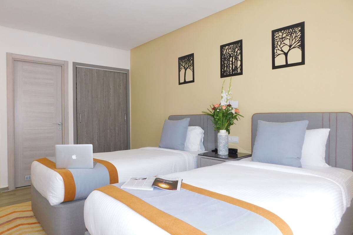 Une chambre d’hôtel avec deux lits, un ordinateur portable posé sur l’un des lits, un vase rempli de fleurs sur une table de chevet, et des œuvres d’art accrochées au mur.