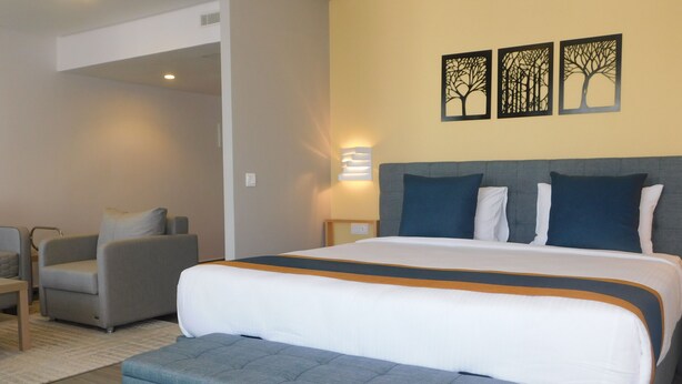 Une chambre d’hôtel avec un grand lit, un banc recouvert d’un tissu gris, un fauteuil gris et une petite table. Trois œuvres d’art sont accrochées au mur et représentent des arbres.
