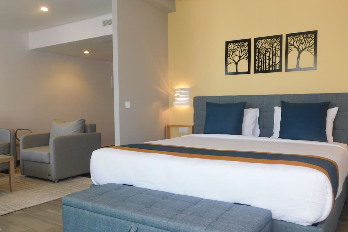 Une chambre d’hôtel avec un grand lit, un banc recouvert d’un tissu gris, un fauteuil gris et une petite table. Trois œuvres d’art sont accrochées au mur et représentent des arbres.