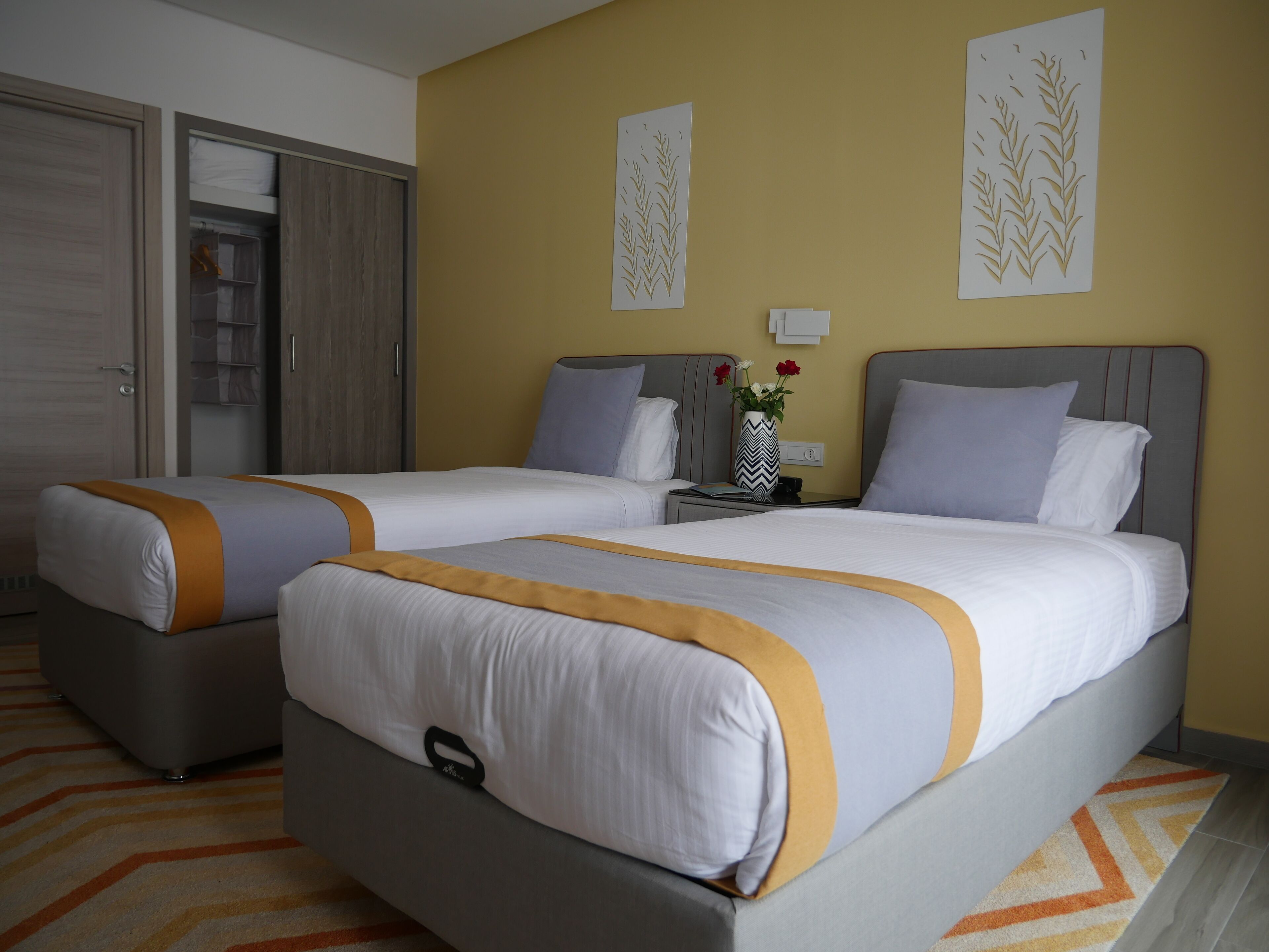 Habitación de hotel con dos camas, un armario, una mesita de noche con un florero y una pared con paneles decorativos.
