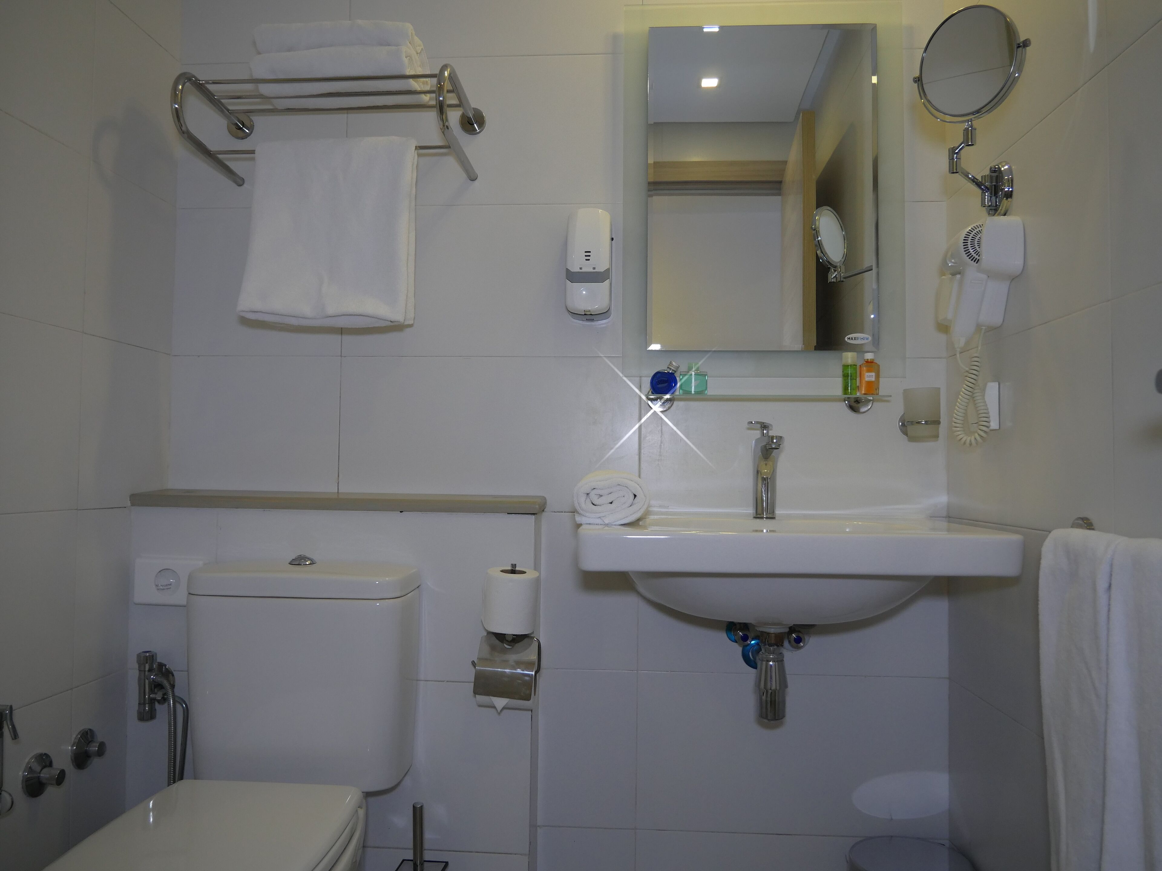 A bathroom with a toilet, sink, and mirror. There is a towel rack above the toilet and a hairdryer on the wall.