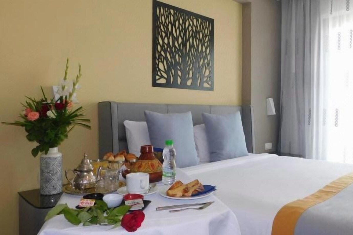 A hotel room with a bed, a table with breakfast items, a vase with flowers, and a decorative wall piece.