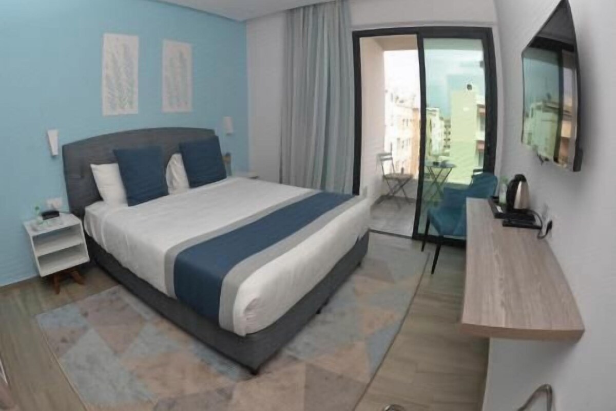 A modern hotel room with a large bed, a desk, a chair, and a balcony with a view.