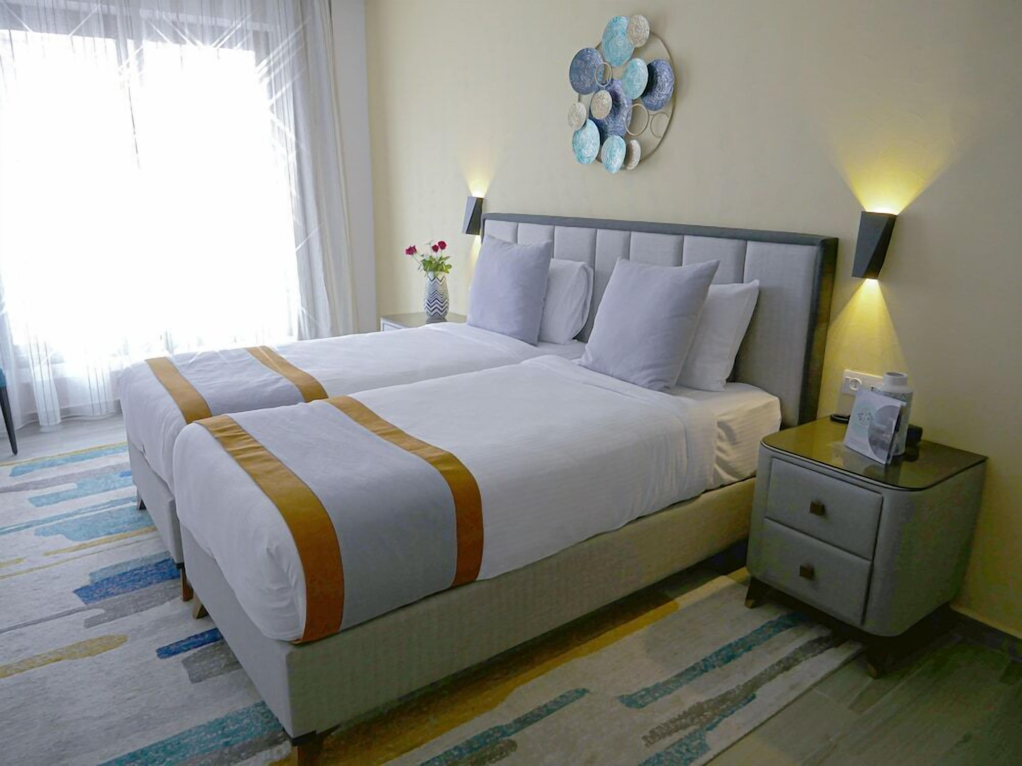Cama doble con ropa blanca y un detalle dorado, una mesita de noche con lámpara y un adorno en la pared.