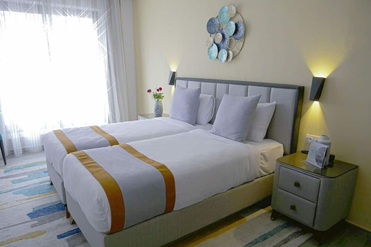 A double bed with white bedding and a gold accent, a bedside table with a lamp, and a decorative wall piece.