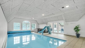 Una piscina interior con un tobogán azul, ventanas amplias y una planta en maceta.