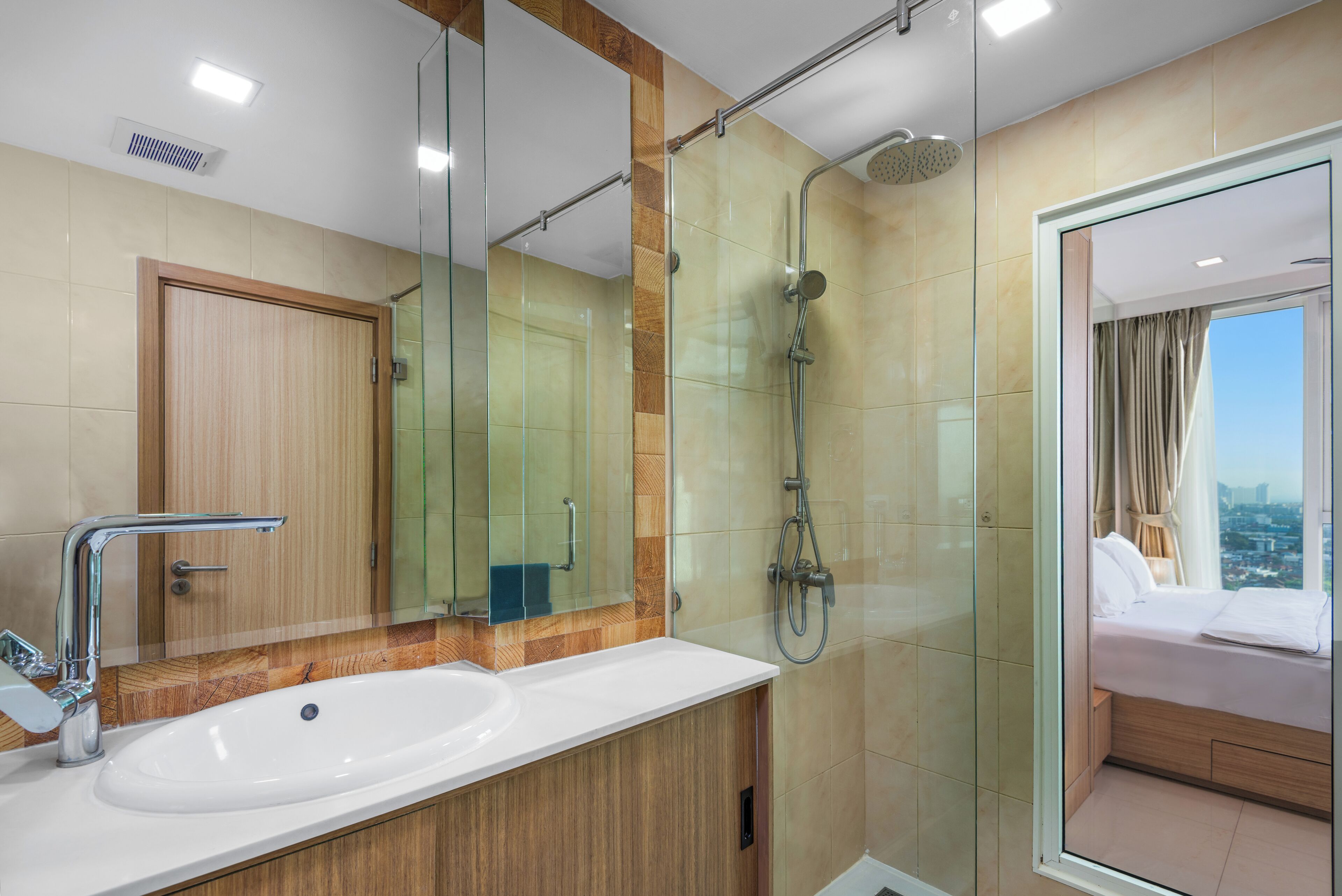 Ein modernes Badezimmer mit Duschkabine aus Glas, einem großen Spiegel und Blick auf ein Schlafzimmer mit Bett und Vorhängen.