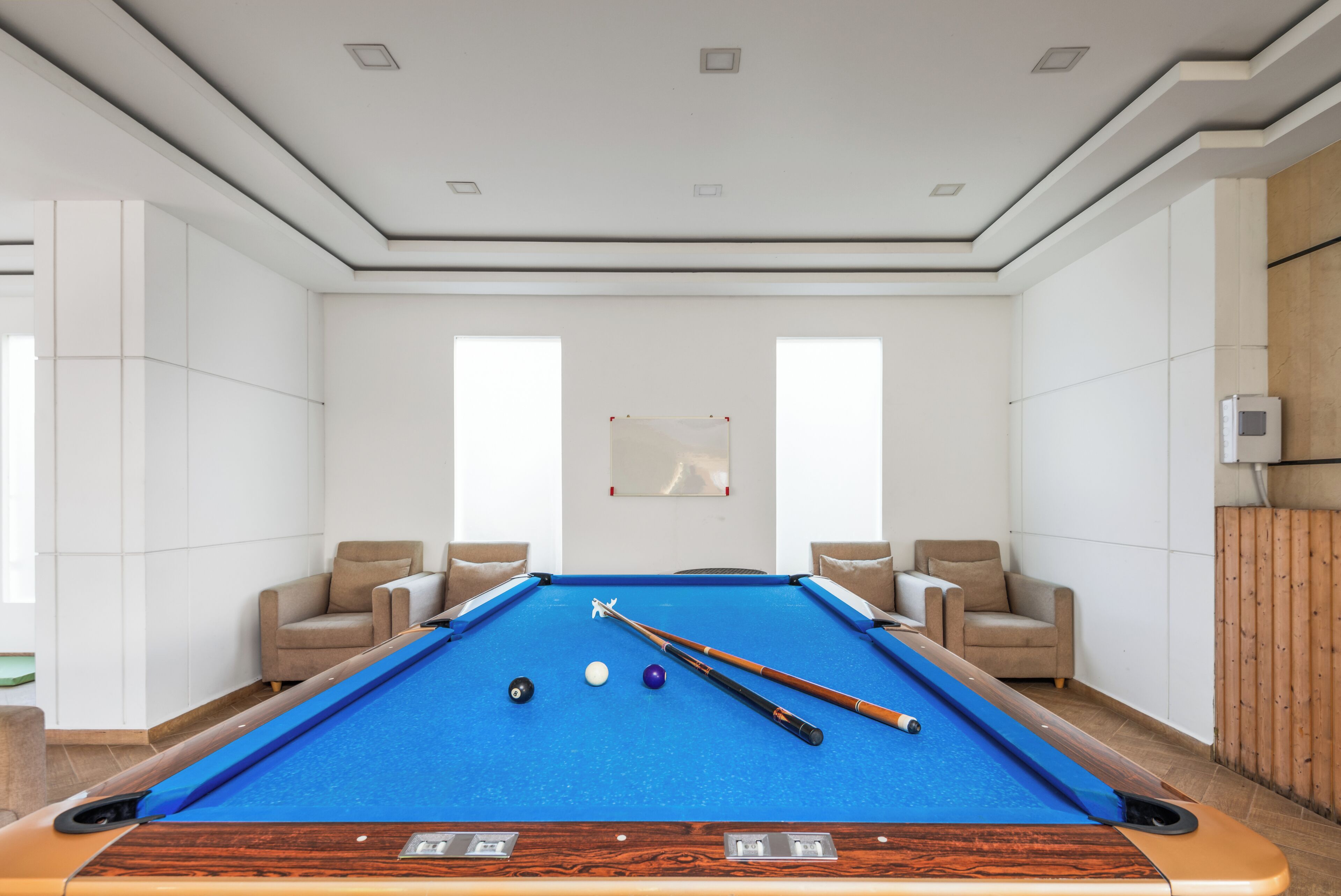 A pool table with balls and cues, surrounded by two armchairs, in a room with white walls and a wooden panel on one side.