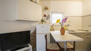 Une cuisine de taille réduite avec des meubles blancs, un réfrigérateur, une table à manger avec des chaises, un téléviseur et une fenêtre avec un rideau.