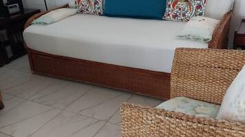 Un letto con un cuscino blu, cuscini decorati e una sedia in vimini con un cuscino floreale.