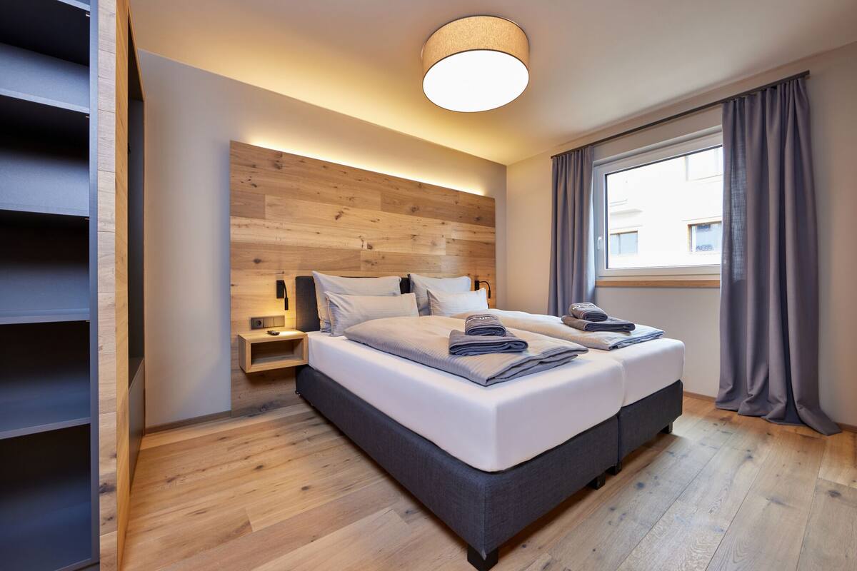 Ein modernes Schlafzimmer mit einem hölzernen Kopfteil, einem großen Bett mit ordentlich gefalteten Handtüchern, einem Nachttisch und einem Fenster mit Vorhängen.