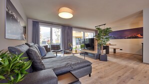 Un salon moderne avec un canapé gris, un coin repas comprenant une table et des chaises, une grande télévision et une vue sur l’extérieur grâce à de grandes fenêtres.