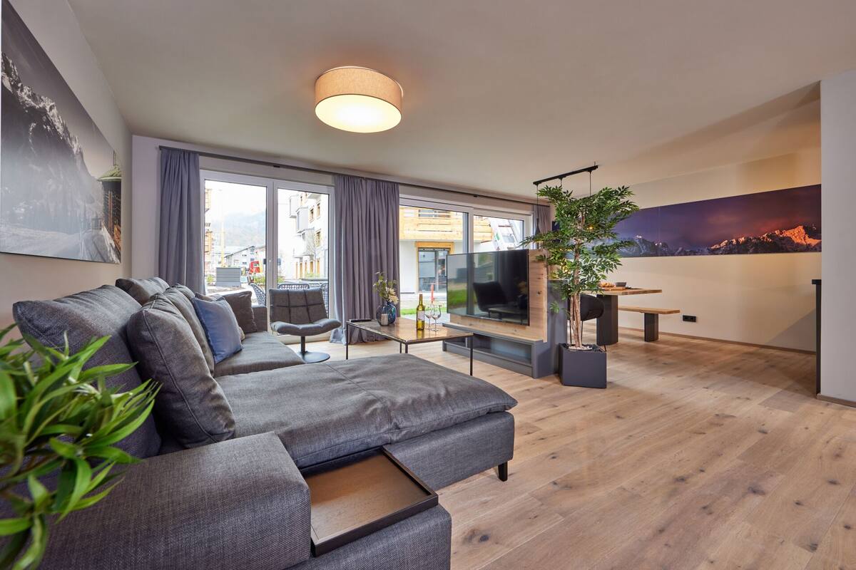 Ein modernes Wohnzimmer mit grauem Sofa, einem Essbereich mit Tisch und Stühlen, einem großen Fernseher und Blick ins Freie durch große Fenster.