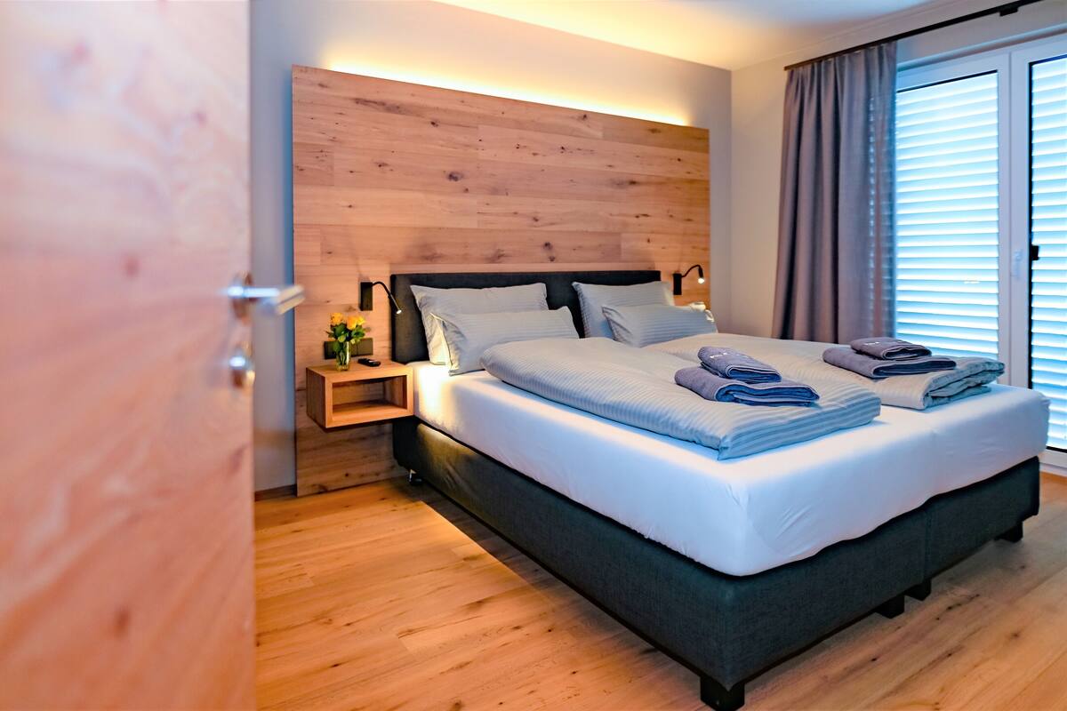 Ein modernes Schlafzimmer mit einem Holz-Kopfteil, einem ordentlich bezogenen Bett mit gefalteten Handtüchern, einem Nachttisch mit Lampe und einem Fenster mit Jalousien.