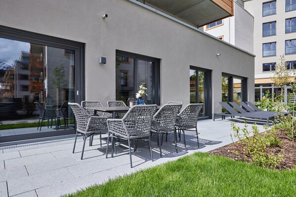 Ein Sitzbereich im Freien mit Korbstühlen und einem Tisch, ein kleiner Garten und ein Gebäude mit großen Fenstern.
