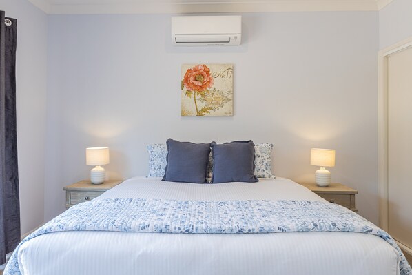 A bedroom with a bed, two bedside tables with lamps, a wall-mounted air conditioner, and a floral artwork above the bed.