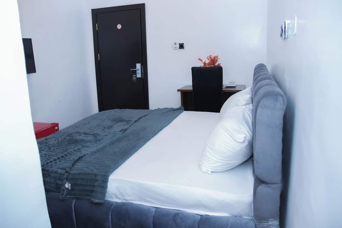 A hotel room with a bed, a desk, a chair, a door, and a small table with a vase of flowers.