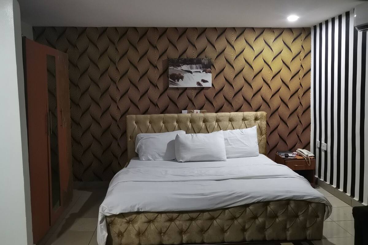 A bedroom with a bed, a bedside table, a nightstand, a patterned wall, and a door.
