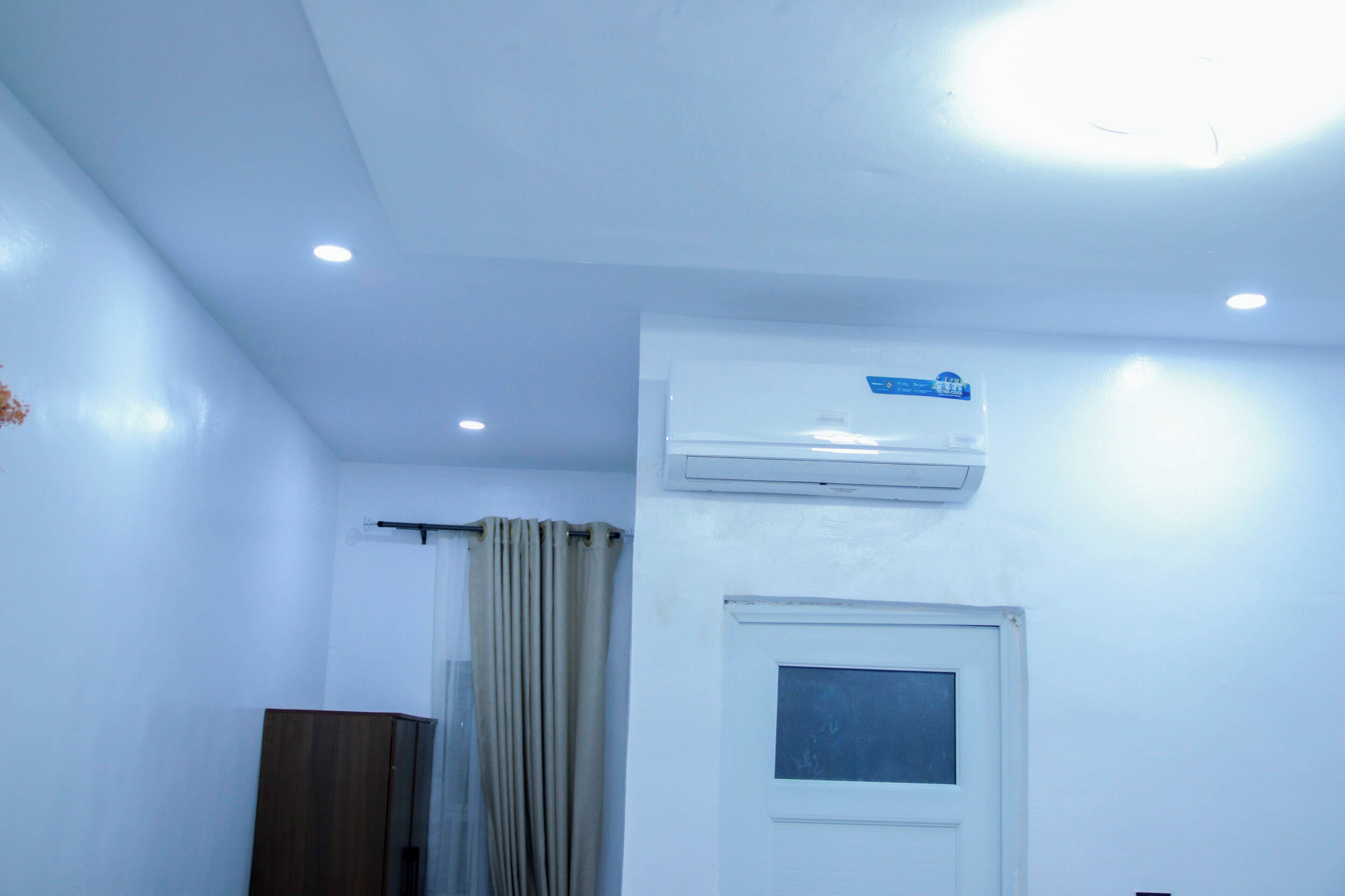 A room with white walls, a window with a curtain, an air conditioning unit mounted above the window, and a door with a small window.