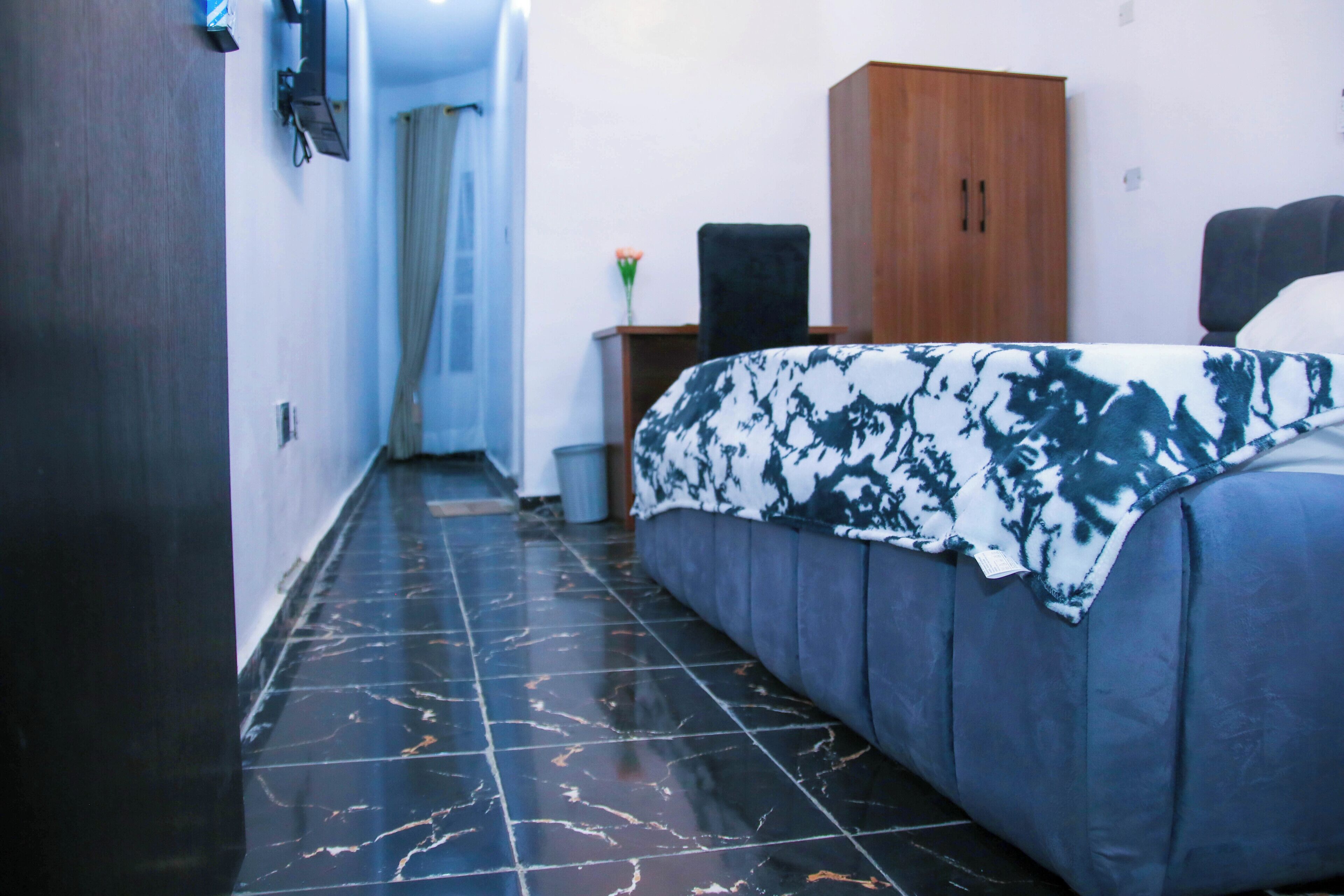 A hotel room with a bed, a wooden wardrobe, a small table with a vase, and a tiled floor.