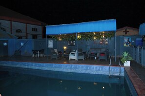Un área de piscina al aire libre con una zona de estar cubierta, una piscina con borde decorativo y un edificio al fondo.