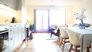 A modern kitchen with white cabinets, a dining area with a table and chairs, and a large window providing natural light. - Central & Cozy - Rainbow A (Lens)