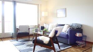 A living room with a sofa, armchair, and a coffee table. There is a window with curtains, a lamp, and a framed picture on the wall. - Central & Cozy - Rainbow A (Lens)