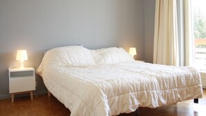 A bedroom with a bed, two bedside tables with lamps, a window with curtains, and a view of greenery outside. - Cozy & Central - Palma B (Lens)