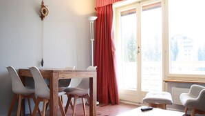 A room with a wooden dining table, four chairs, a small table with a remote control, and a window with red curtains. - Cozy & Central - Palma B (Lens)