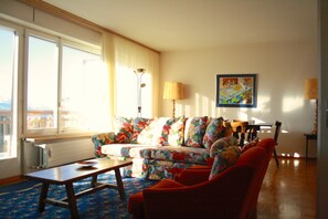 A living room with a sofa, a coffee table, and a chair. There is a window with curtains, a lamp, and a painting on the wall. - Mountain View & Spacious - Flaminia C (Lens)
