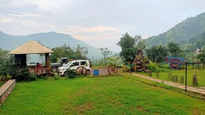 A rural setting with a white car parked near a wooden structure with a thatched roof, surrounded by greenery and hills in the background. - I-Camp Resort (Wai)